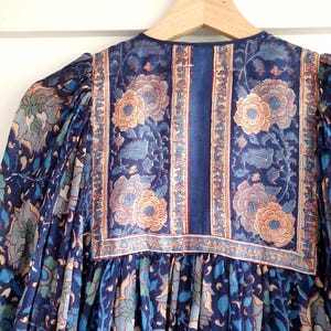 May include: A blue floral print dress with long sleeves hangs on a wooden hanger. The dress features a detailed pattern of flowers and leaves in shades of blue, teal, and peach. The neckline and yoke are adorned with a contrasting floral border.