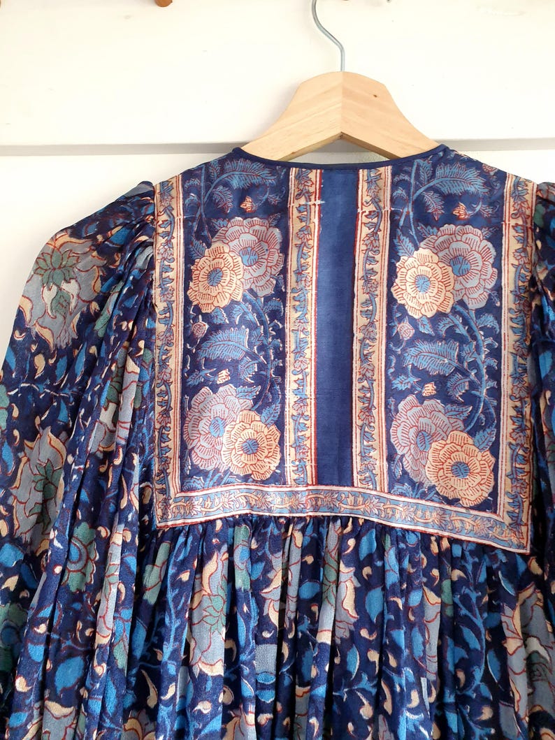 May include: A blue floral print dress with long sleeves hangs on a wooden hanger. The dress features a detailed pattern of flowers and leaves in shades of blue, teal, and peach. The neckline and yoke are adorned with a contrasting floral border.