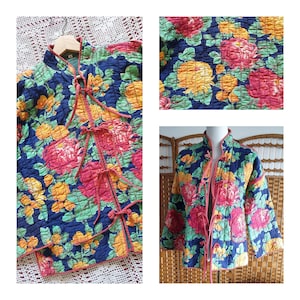 Puede incluir: Chaqueta acolchada con un vibrante estampado floral en tonos rosa, amarillo y verde sobre un fondo azul marino. La chaqueta presenta un cuello mandarín y cierres de lazo. La chaqueta se muestra en una percha y un maniquí.