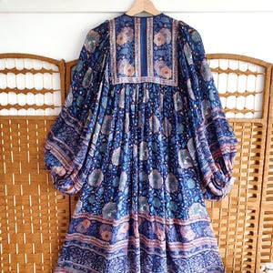 May include: A blue floral print dress with long, voluminous sleeves hangs on a wooden hanger. The dress features a V-neck and a gathered waist. The fabric has a detailed pattern with shades of blue, pink, and cream. The dress is displayed against a wooden screen.