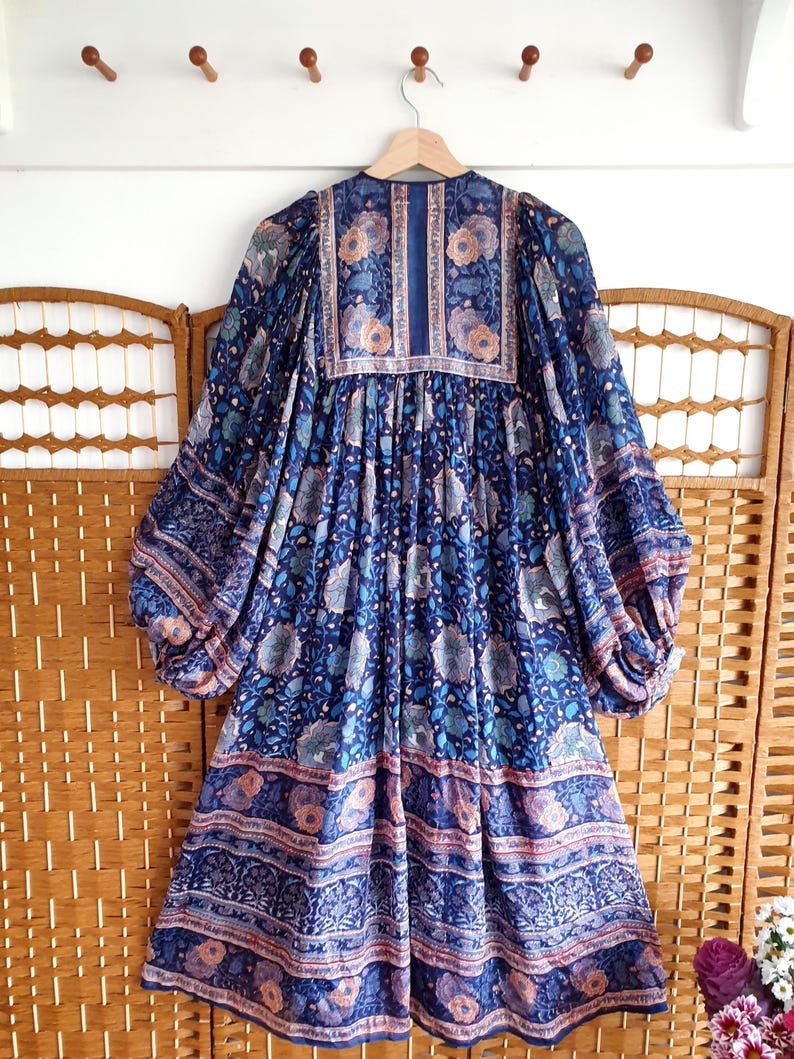 May include: A blue floral print dress with long, voluminous sleeves hangs on a wooden hanger. The dress features a V-neck and a gathered waist. The fabric has a detailed pattern with shades of blue, pink, and cream. The dress is displayed against a wooden screen.