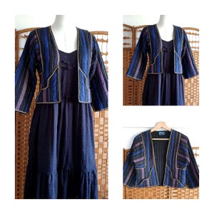 Puede incluir: Un vestido azul marino con una chaqueta a rayas a juego. La chaqueta presenta rayas verticales en tonos azules, morados y negros, delineadas con ribetes dorados. El vestido tiene una falda escalonada y un escote redondo. El conjunto se exhibe en un maniquí.