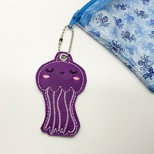 May include: Purple felt jellyfish keychain with a smiling face and pink eyelashes. The keychain is attached to a silver chain.