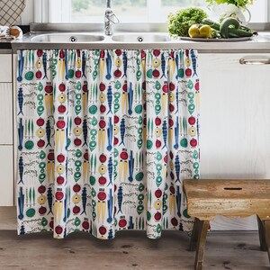 Może przedstawiać: Biały zlew kuchenny ze stalową misą i białym blatem. Pod zlewozmywakiem wisi kolorowa zasłona z tkaniny z wzorem ryb, marchewek i pomidorów. Przed zlewozmywakiem stoi drewniany stołek.