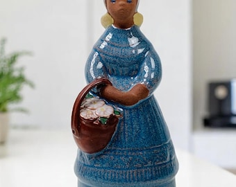 May include: A ceramic figurine of a woman in a blue dress holding a brown basket with white flowers. The woman has blonde hair and a blue headscarf.
