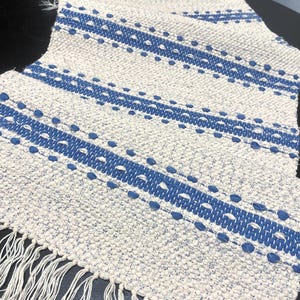 Puede incluir: Un camino de mesa tejido en blanco roto y azul con borde de flecos. El camino de mesa presenta rayas azules y detalles punteados. El textil está hecho de un material texturizado.