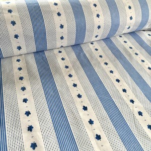 Può includere: Un rotolo di tessuto con un motivo ripetuto di strisce blu e motivi a foglie blu su sfondo bianco. Il tessuto è arrotolato, rivelando il motivo. Le strisce alternano tra linee piene e tratteggiate.