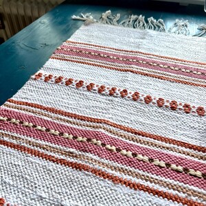 Peut inclure: Un tapis rectangulaire tissé avec une base blanche et des rayures horizontales dans des tons de rose, de marron et de beige. Le tapis présente un motif texturé avec des franges à une extrémité. Le tapis est posé sur une surface turquoise.