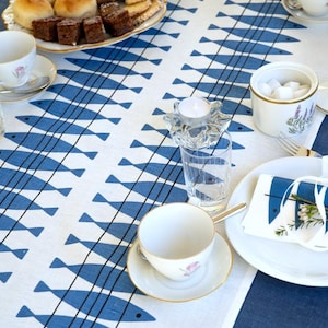 Può includere: Una tavola apparecchiata con una tovaglia bianca con un motivo a pesci blu. Tazze da tè, piattini e piatti sono disposti, insieme a un piatto di pasticcini. Un piccolo portacandele in vetro completa la scena.