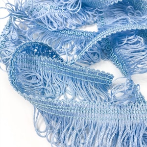 May include: Light blue fringe trim with a delicate, looped texture. The trim is made of soft, woven fabric and has a subtle sheen.