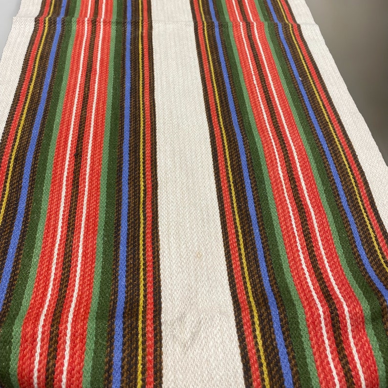 May include: A white woven fabric with red, green, blue, and brown stripes. The fabric has a textured surface and is perfect for a variety of crafting projects.