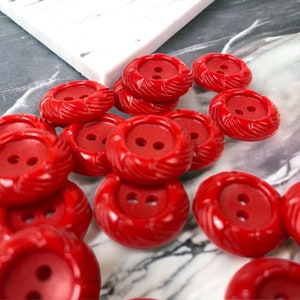 May include: A pile of red plastic buttons with two holes each. The buttons have a raised, textured design around the edges.
