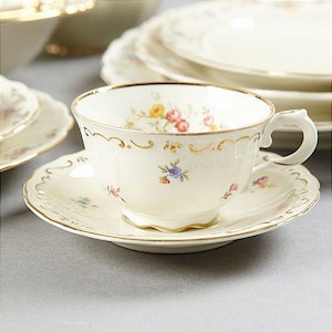 Könnte beinhalten: Cremefarbenes Tee-Set mit floralen und goldenen Verzierungen. Die Teetasse hat einen zarten Henkel und steht auf einer passenden Untertasse. Weitere Teller sind im Hintergrund gestapelt, was auf ein komplettes Teeservice hindeutet.
