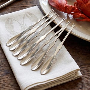 May include: A set of six silver seafood forks arranged on a folded white linen napkin with a subtle embossed design. A cooked lobster rests on a white plate, set on a dark wooden table. Additional silverware is visible.