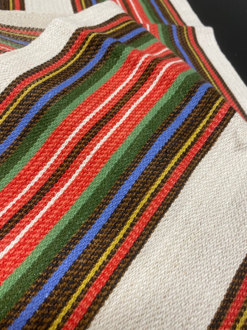 May include: A white fabric with multi-colored stripes in red, green, blue, brown, and yellow. The fabric is woven with a textured pattern.