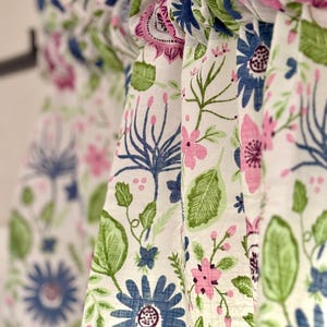 May include: Close-up of floral patterned curtains. The curtains feature a white background with a repeating pattern of blue, pink, and green flowers and leaves. The fabric is gathered at the top, creating soft folds.