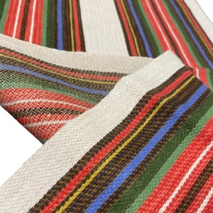 May include: A close-up of a woven fabric with red, green, white, blue, and brown stripes. The fabric has a subtle shimmer and is likely used for a tablecloth or other home decor.