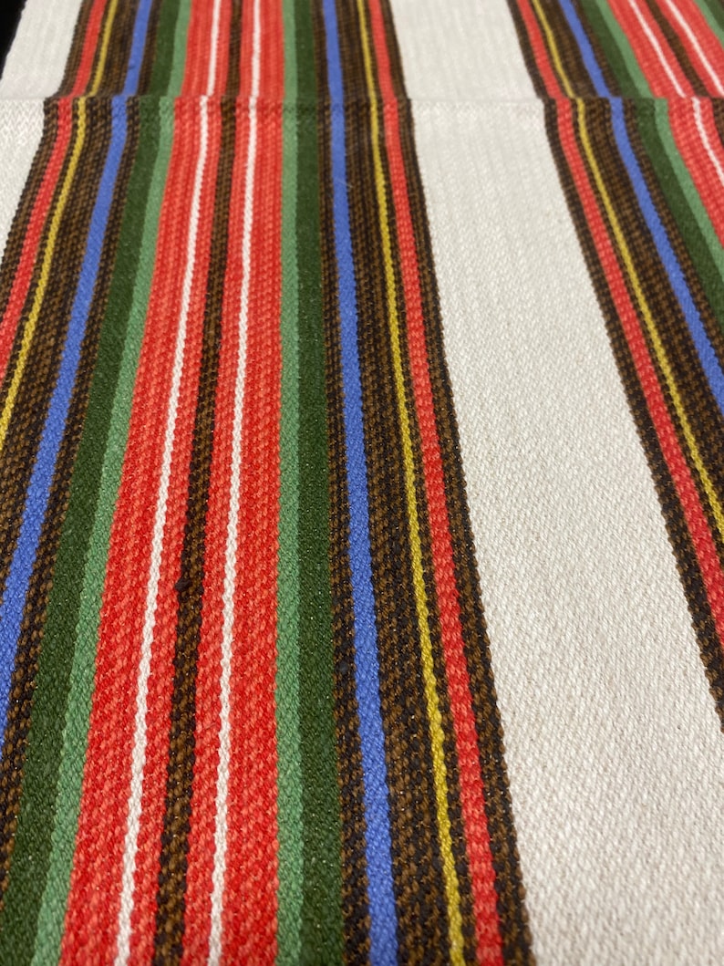 May include: A white fabric with red, green, blue, brown, and yellow stripes. The fabric has a woven texture.