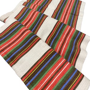May include: A white fabric runner with red, green, blue, brown, and yellow stripes. The runner is folded in thirds and has a woven texture.