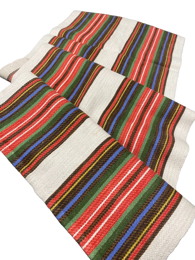 May include: A white fabric runner with red, green, blue, brown, and yellow stripes. The runner is folded in thirds and has a woven texture.