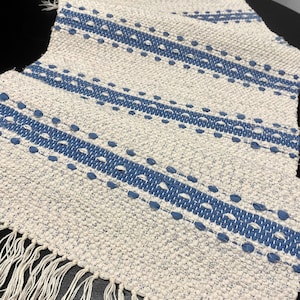 Può includere: Un tappeto tessuto bianco e blu con frange sui bordi. Il tappeto ha un motivo a strisce con strisce blu e punti bianchi.