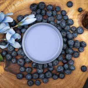 May include: A light blue ceramic bowl with a white rim sits on a wooden surface surrounded by blueberries.  A sprig of blue flowers with green leaves is on the left side of the bowl.