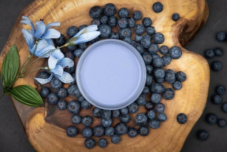 May include: A light blue ceramic bowl with a white rim sits on a wooden surface surrounded by blueberries.  A sprig of blue flowers with green leaves is on the left side of the bowl.