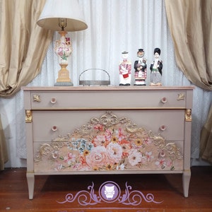 May include: A light pink dresser with three drawers and gold accents. The top drawer has a floral design with pink, white, and yellow flowers. The dresser is on a wooden floor. The Fancy Purple House logo is in the bottom right corner.