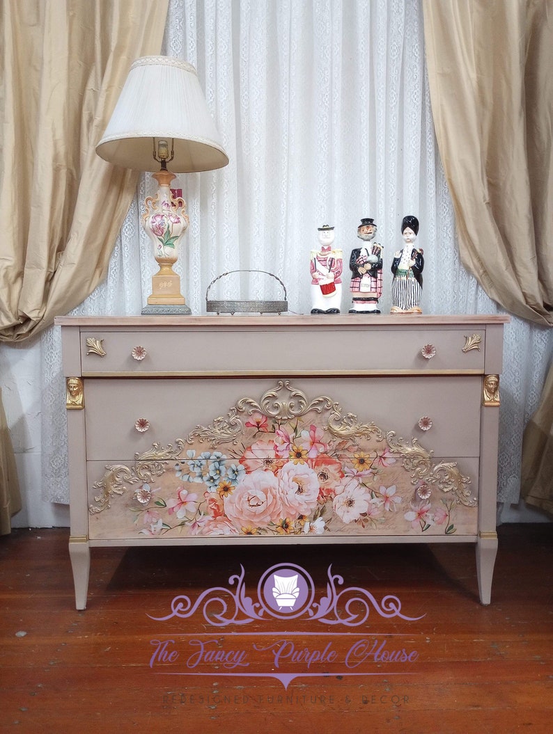 May include: A light pink dresser with three drawers and gold accents. The top drawer has a floral design with pink, white, and yellow flowers. The dresser is on a wooden floor. The Fancy Purple House logo is in the bottom right corner.