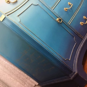 May include: A blue and navy blue dresser with gold accents and ornate hardware. The dresser has a gradient paint finish, transitioning from a lighter blue to a darker navy blue.