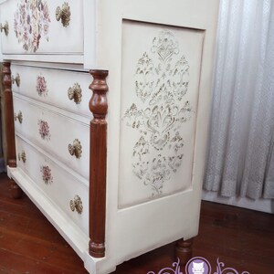 May include: A white dresser with four drawers and brown wooden legs. The dresser has a decorative stencil design on the front and floral designs on the drawers. The dresser is painted in a distressed white finish.