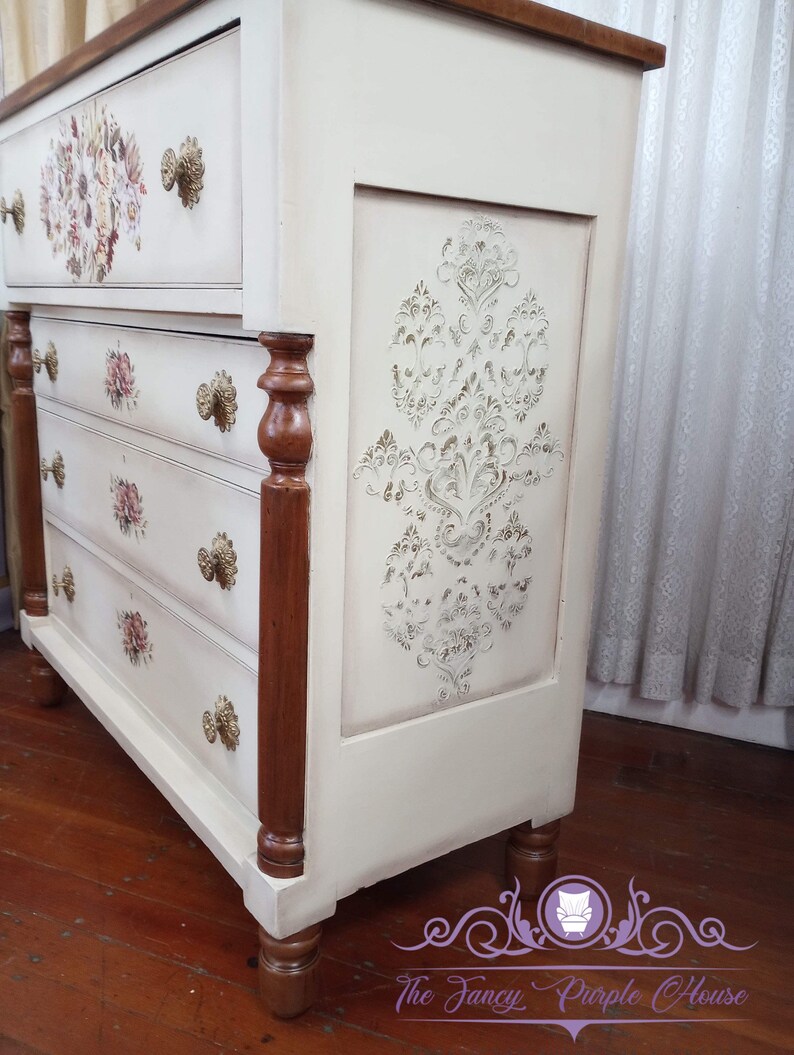 May include: A white dresser with four drawers and brown wooden legs. The dresser has a decorative stencil design on the front and floral designs on the drawers. The dresser is painted in a distressed white finish.