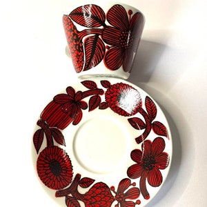 Pode incluir: Uma ch&aacute;vena de caf&eacute; e um pires de cer&acirc;mica branca com um padr&atilde;o floral vermelho e preto. A ch&aacute;vena e o pires s&atilde;o decorados com flores e folhas estilizadas.