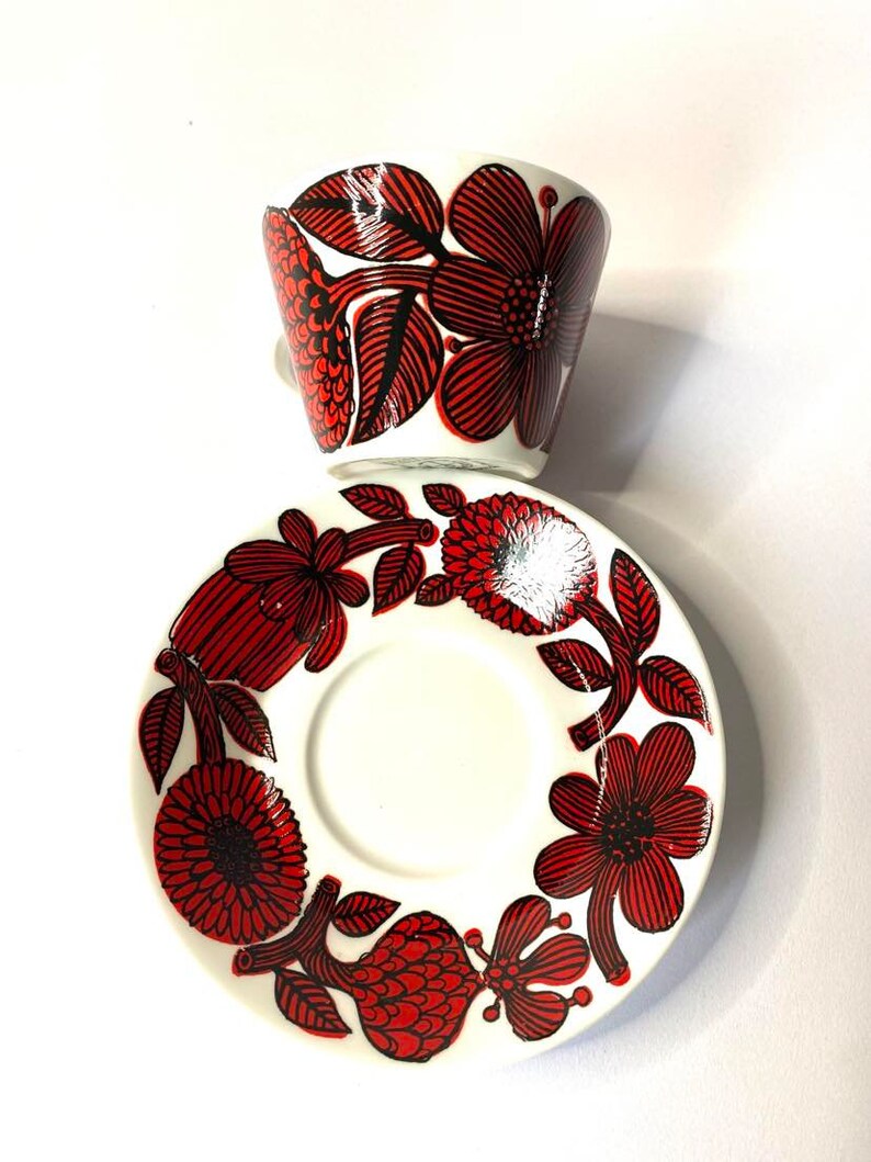 Pode incluir: Uma ch&aacute;vena de caf&eacute; e um pires de cer&acirc;mica branca com um padr&atilde;o floral vermelho e preto. A ch&aacute;vena e o pires s&atilde;o decorados com flores e folhas estilizadas.
