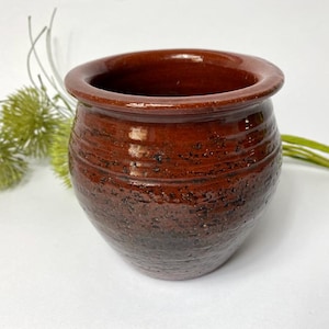 Puede incluir: Una pequeña maceta de cerámica marrón con una superficie texturizada. La maceta tiene una forma redondeada y una abertura ancha.
