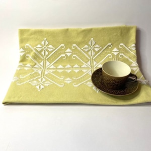Op de afbeelding: Een gele geborduurde theedoek met een wit geometrisch patroon en een bruin theekopje en schoteltje erop.