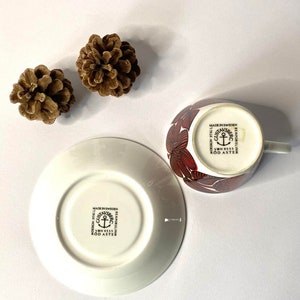 Pode incluir: Uma ch&aacute;vena de ch&aacute; e um pires brancos com um padr&atilde;o floral vermelho. O pires tem o texto "MADE IN SWEDEN DESIGN STIGLI GUSTAVSBERG BENPORSLING VBN 8555 ROD ASTER" impresso na parte inferior.