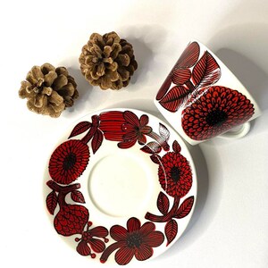 Pode incluir: Uma ch&aacute;vena de ch&aacute; e um pires de cer&acirc;mica branca com um padr&atilde;o floral vermelho e preto. A ch&aacute;vena e o pires t&ecirc;m um design floral estilizado com flores vermelhas e contornos pretos.