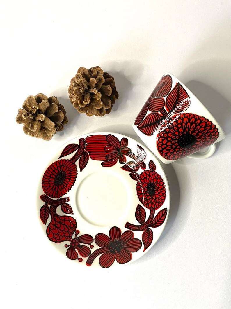Pode incluir: Uma ch&aacute;vena de ch&aacute; e um pires de cer&acirc;mica branca com um padr&atilde;o floral vermelho e preto. A ch&aacute;vena e o pires t&ecirc;m um design floral estilizado com flores vermelhas e contornos pretos.
