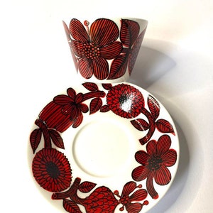 Pode incluir: Um conjunto de ch&aacute;vena e pires de caf&eacute; de cer&acirc;mica branca com um padr&atilde;o floral vermelho e preto. A ch&aacute;vena e o pires t&ecirc;m um design floral estilizado com flores e folhas vermelhas e pretas.