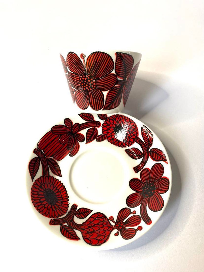Pode incluir: Um conjunto de ch&aacute;vena e pires de caf&eacute; de cer&acirc;mica branca com um padr&atilde;o floral vermelho e preto. A ch&aacute;vena e o pires t&ecirc;m um design floral estilizado com flores e folhas vermelhas e pretas.