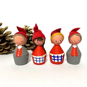 Op de afbeelding: Vier houten beeldjes van gnomen met rode mutsen en beschilderde gezichten. De gnomen dragen verschillende outfits, waaronder een rood-wit geruite jurk, een rood-wit gestreepte jurk en een grijs pak.