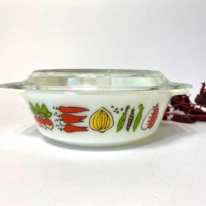 May include: White ceramic casserole dish with a glass lid. The dish has a colorful design of vegetables, including carrots, onions, peas, and a red pepper.
