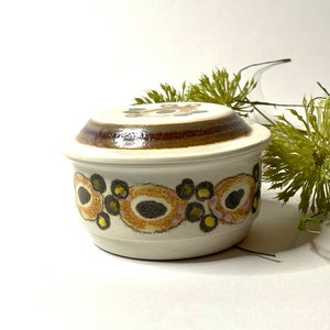 Può includere: Un barattolo di ceramica bianca con un coperchio. Il barattolo ha un motivo floreale marrone, giallo e nero intorno al centro. Il coperchio ha un motivo floreale marrone e giallo.