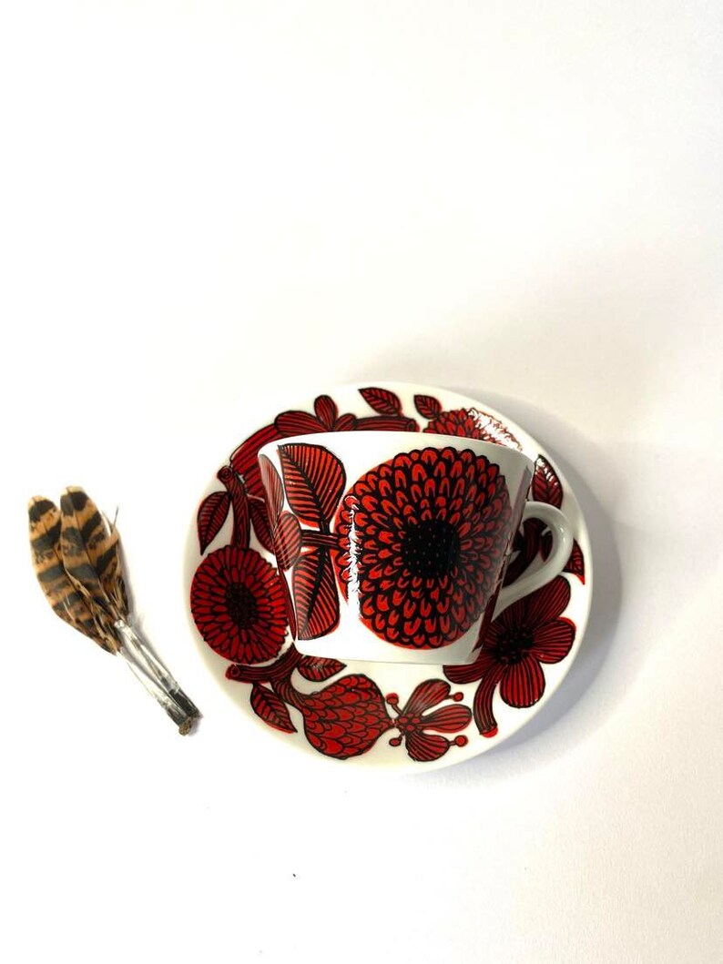 Pode incluir: Uma ch&aacute;vena de ch&aacute; e um pires de cer&acirc;mica branca com um padr&atilde;o floral vermelho e preto. A ch&aacute;vena e o pires s&atilde;o decorados com flores e folhas estilizadas.