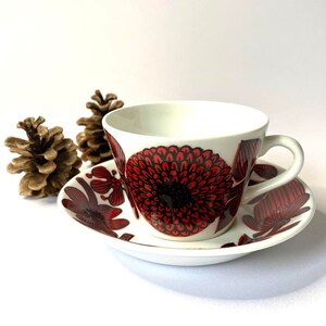 Pode incluir: Uma ch&aacute;vena de ch&aacute; e um pires brancos com um padr&atilde;o floral vermelho e preto. A ch&aacute;vena e o pires est&atilde;o sobre uma superf&iacute;cie branca.
