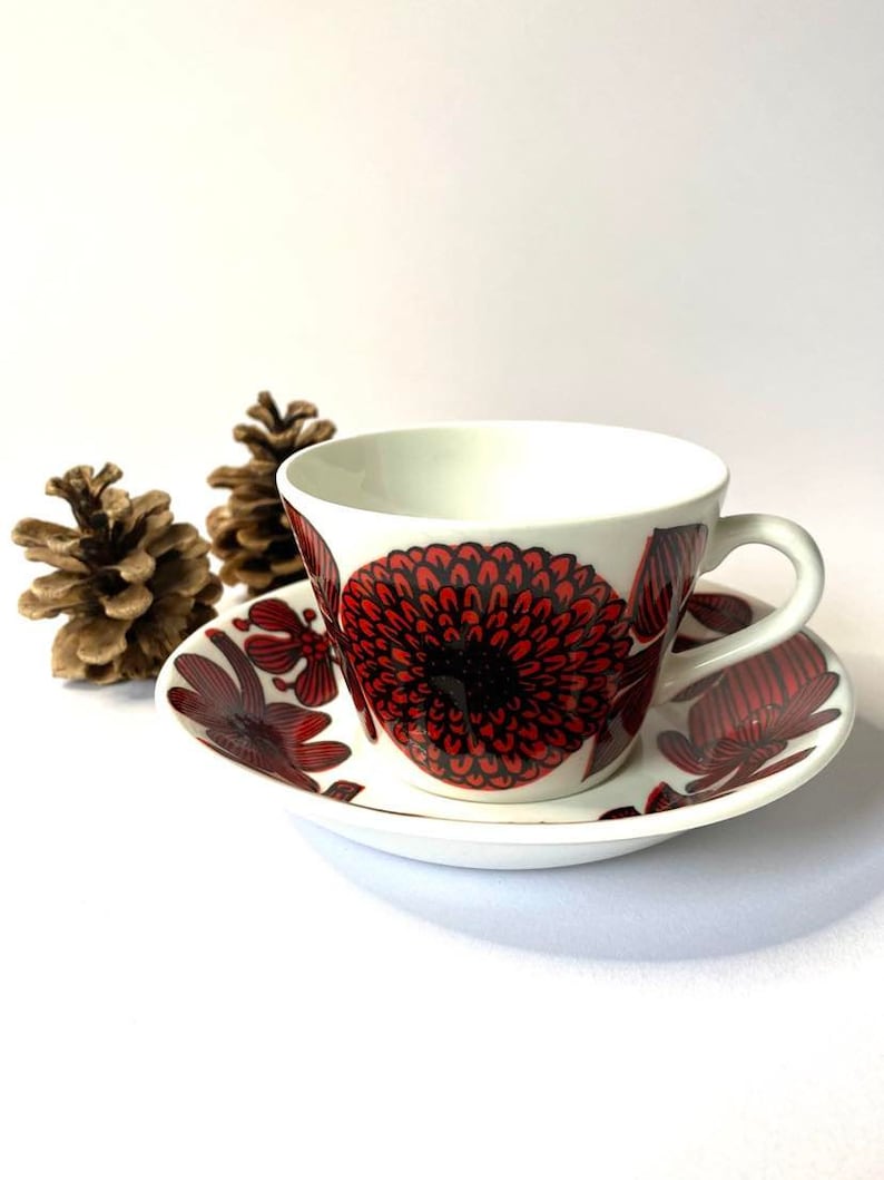 Pode incluir: Uma ch&aacute;vena de ch&aacute; e um pires brancos com um padr&atilde;o floral vermelho e preto. A ch&aacute;vena e o pires est&atilde;o sobre uma superf&iacute;cie branca.
