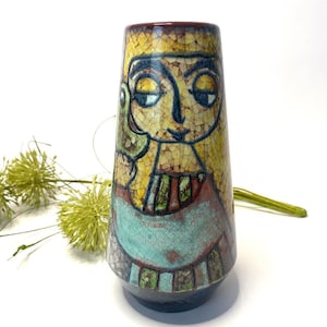 May include: A ceramic vase with a yellow, blue, green, and brown glaze. The vase features a stylized illustration of a person with a bird on their shoulder.