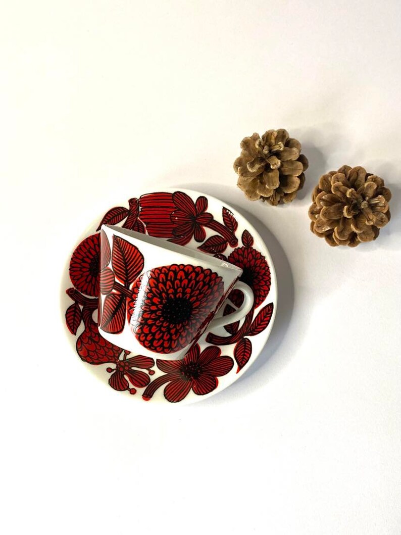 Pode incluir: Uma ch&aacute;vena de ch&aacute; e um pires de cer&acirc;mica branca com um padr&atilde;o floral vermelho e preto. A ch&aacute;vena est&aacute; inclinada para o lado e o pires est&aacute; por baixo.