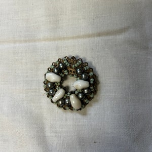 Puede incluir: Un broche circular hecho de cuentas plateadas y blancas. El broche tiene la forma de una corona con una abertura central.